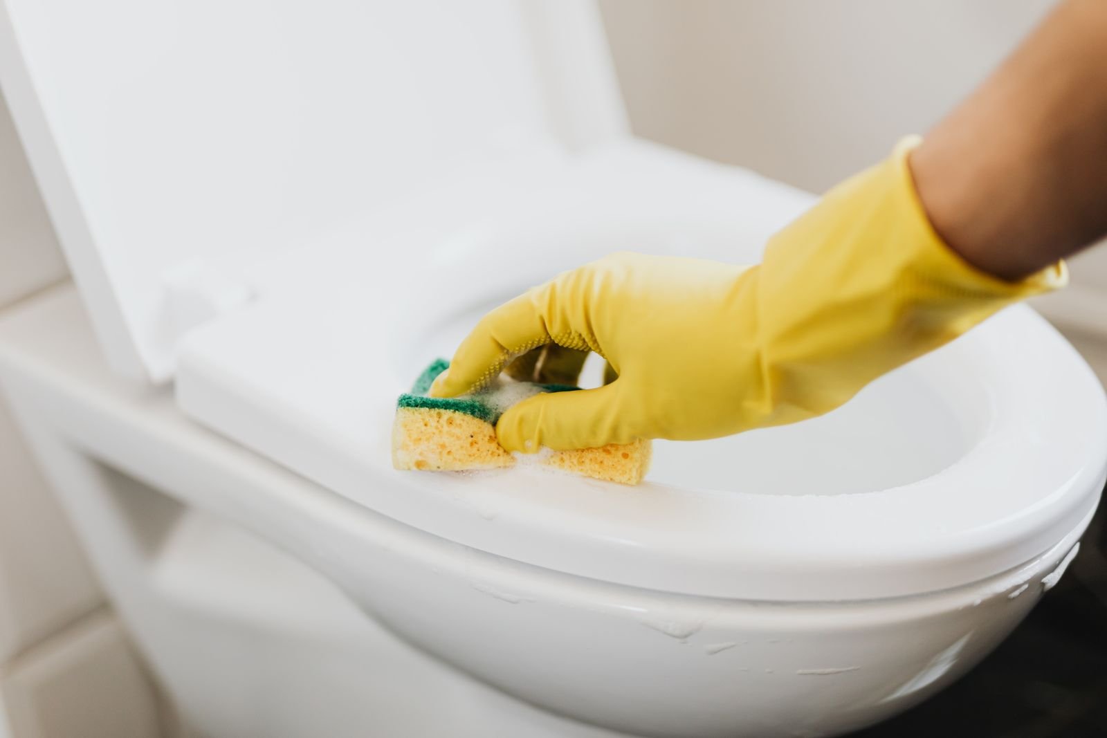 A hand in a yellow rubber glove cleaning a toilet seat with a sponge