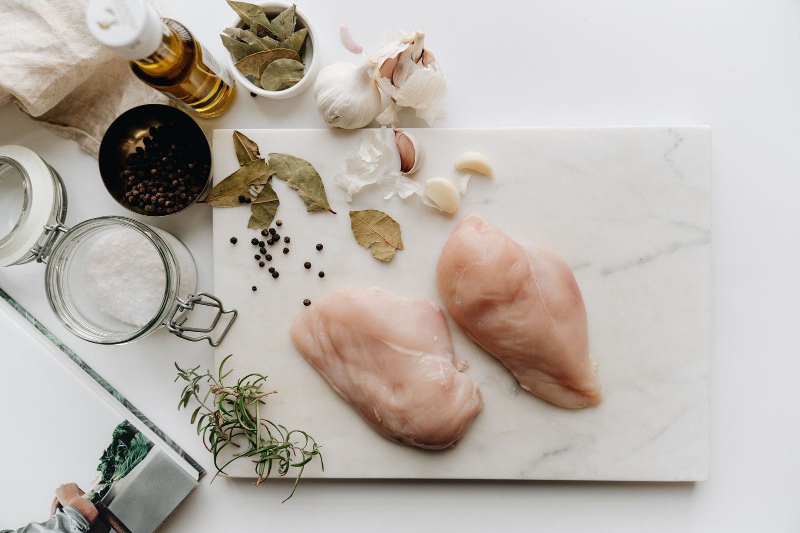 Two raw chicken breasts on a white chopping board with herbs and spices